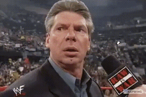 A Man In A Suit And Tie Is Talking Into A Microphone While Wearing A Wwe Logo On His Shirt . GIF