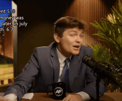 A Man In A Suit And Tie Is Talking Into A Microphone With A Mug That Says Af On It GIF