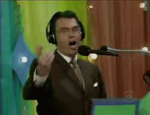 A Man In A Suit And Tie Is Talking Into A Microphone With Headphones On . GIF