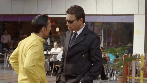 A Man In A Suit And Tie Is Talking To A Man In A Yellow Shirt GIF