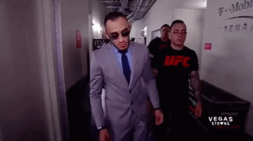 A Man In A Suit And Tie Is Walking Down A Hallway Next To A Man Wearing A Ufc Shirt . GIF