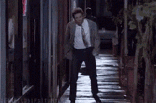 A Man In A Suit And Tie Is Walking Down A Narrow Alleyway At Night . GIF