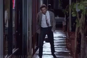 A Man In A Suit And Tie Is Walking Down A Wet Sidewalk At Night . GIF