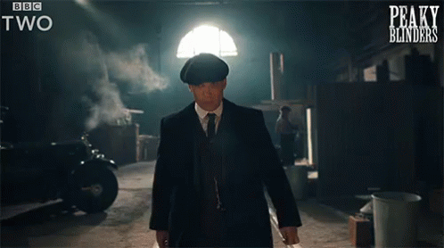 A Man In A Suit And Tie Is Walking In A Dark Room With Peaky Blinders Written On The Bottom Of The Screen GIF