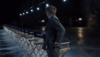 A Man In A Suit And Tie Is Walking In Front Of A Row Of Chairs GIF