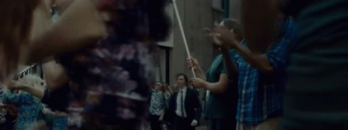 A Man In A Suit And Tie Is Walking Through A Crowd Of People Holding A Stick . GIF