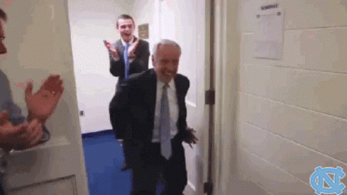 a man in a suit and tie is walking through a doorway while another man applauds .