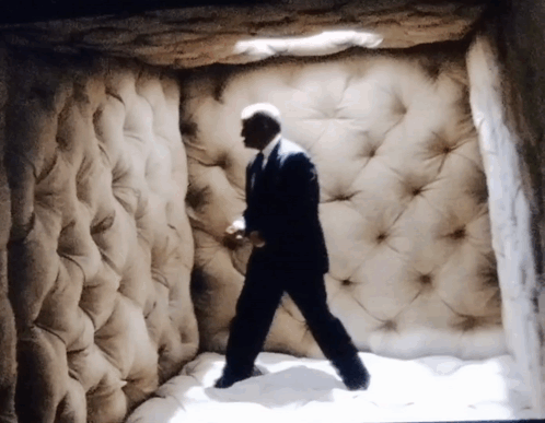 A Man In A Suit And Tie Is Walking Through A Room With A Quilted Wall GIF