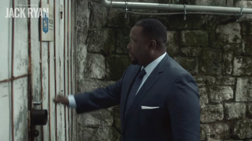 A Man In A Suit And Tie Opens A Door With The Name Jack Ryan On The Wall Behind Him GIF
