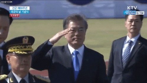A Man In A Suit And Tie Salutes In Front Of A Live Ktv Screen GIF
