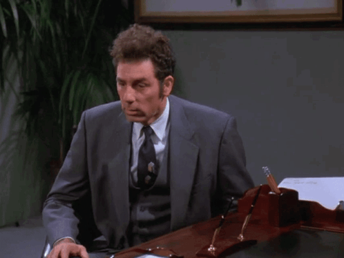 A Man In A Suit And Tie Sits At A Desk With The Words " I Don 'T Even Really Work Here " Above Him GIF