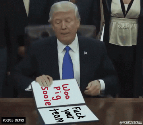 A Man In A Suit And Tie Sits At A Table Holding A Sign That Says " Whoo Pig " GIF