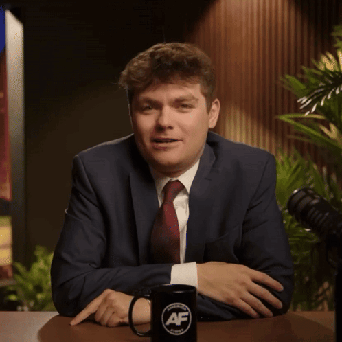 A Man In A Suit And Tie Sits At A Table Next To A Mug That Says Af On It GIF