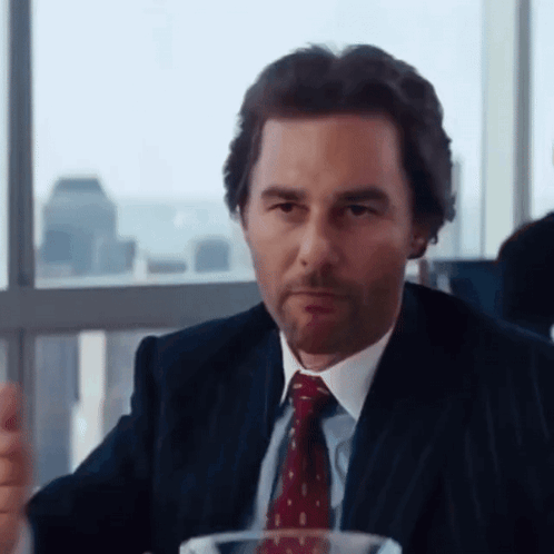 A Man In A Suit And Tie Sits At A Table GIF