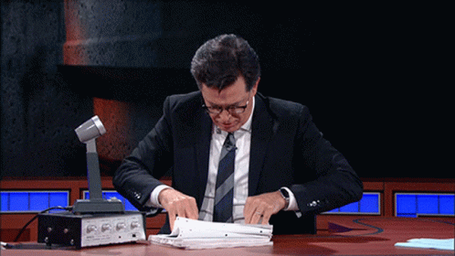 A Man In A Suit And Tie Sits At A Table With A Microphone GIF