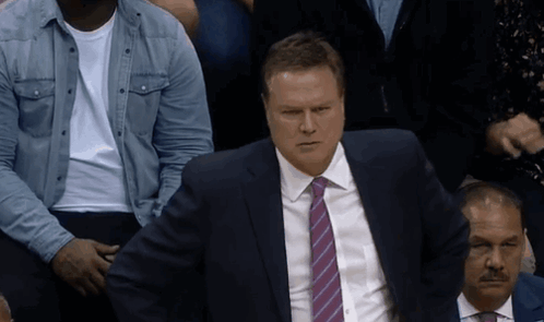 A Man In A Suit And Tie Sits In A Crowd Of People GIF