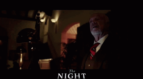 A Man In A Suit And Tie Sits In A Dark Room With The Words The Night Written Above Him GIF