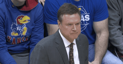 A Man In A Suit And Tie Sits In Front Of A Kansas Sweatshirt GIF