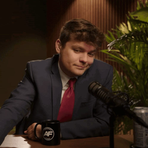 A Man In A Suit And Tie Sits In Front Of A Microphone With A Mug That Says Af GIF