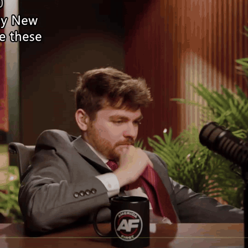 A Man In A Suit And Tie Sits In Front Of A Microphone With A Mug That Says " America First " On It GIF