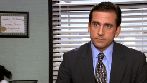A Man In A Suit And Tie Sits In Front Of A Window With Blinds And A Certificate Of Training GIF