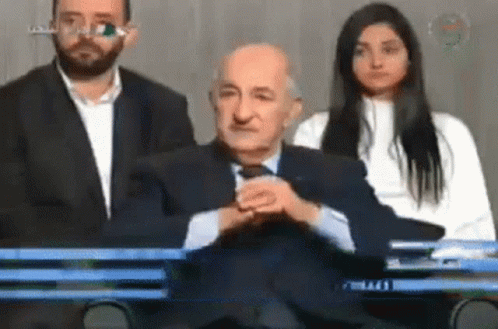 A Man In A Suit And Tie Sits In Front Of A Woman And A Man In A Suit GIF