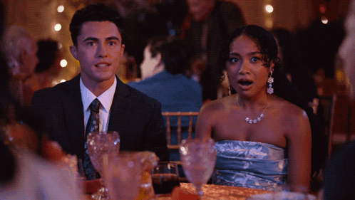A Man In A Suit And Tie Sits Next To A Woman In A Strapless Blue Dress GIF