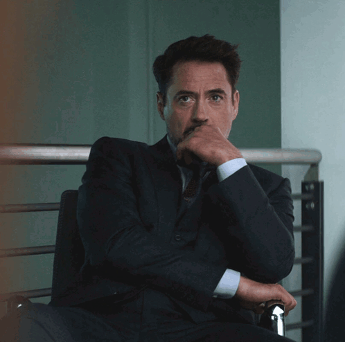 A Man In A Suit And Tie Sits With His Hand On His Chin GIF