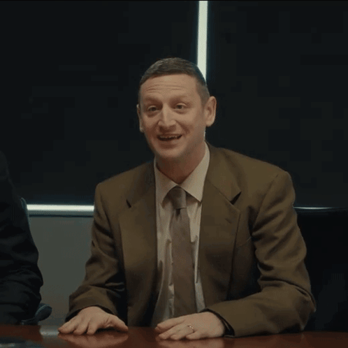 A Man In A Suit And Tie Sitting At A Table With The Word Coward Written On His Face GIF