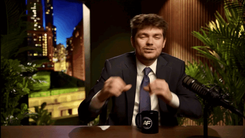 A Man In A Suit And Tie Sitting In Front Of A Microphone With A Mug That Says Af GIF