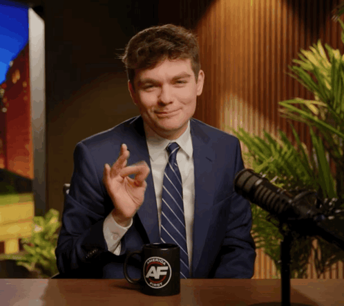 A Man In A Suit And Tie Sitting In Front Of A Microphone With A Mug That Says Avenue Af On It GIF