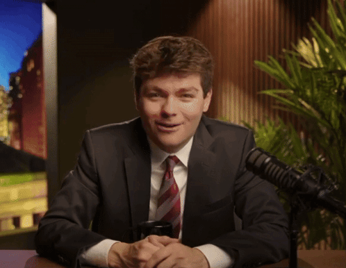 A Man In A Suit And Tie Smiles In Front Of A Microphone GIF