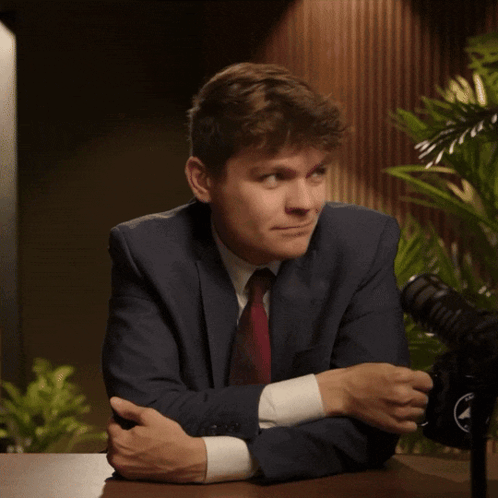 A Man In A Suit And Tie Smiles While Sitting At A Table With A Microphone In The Background GIF