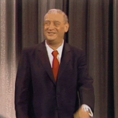 A Man In A Suit And Tie Smiles While Standing In Front Of A Curtain GIF