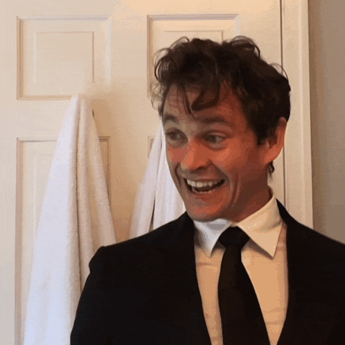 A Man In A Suit And Tie Smiles While Standing In Front Of A Door GIF