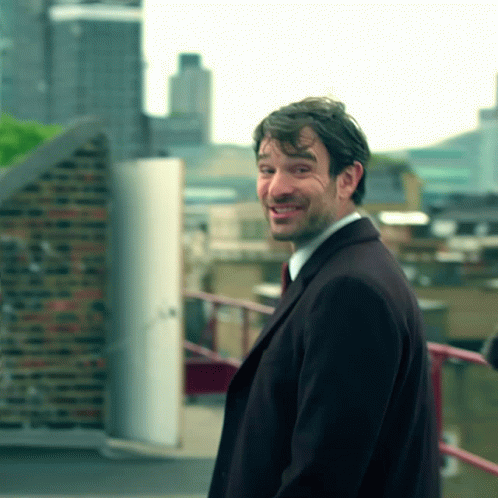 A Man In A Suit And Tie Smiles While Standing On A Balcony GIF