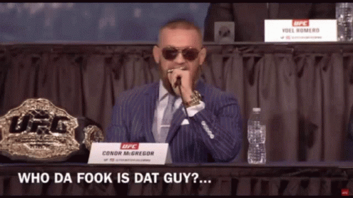 A Man In A Suit And Tie Speaking Into A Microphone With A Sign That Says Conor Mcgregor On It GIF