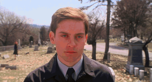 A Man In A Suit And Tie Stands In A Cemetery Looking At The Camera GIF