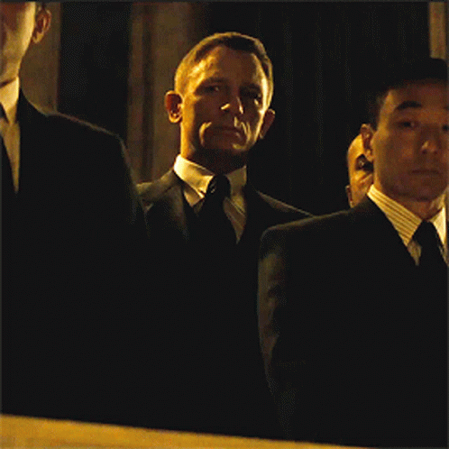 A Man In A Suit And Tie Stands In A Dark Room With Other Men GIF