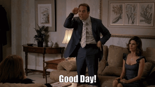 A Man In A Suit And Tie Stands In A Living Room And Says Good Day GIF