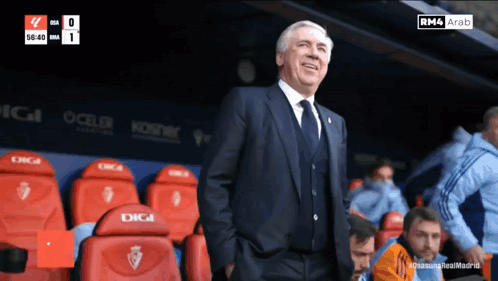 A Man In A Suit And Tie Stands In A Stadium With Red Seats That Say Digi GIF