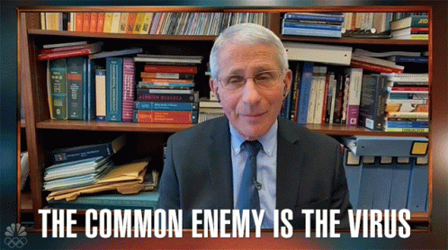 A Man In A Suit And Tie Stands In Front Of A Bookshelf With The Words " The Common Enemy Is The Virus " GIF