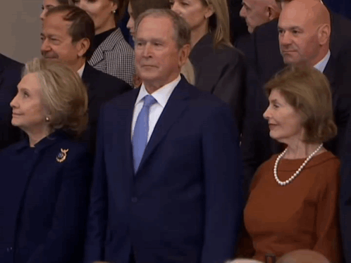 A Man In A Suit And Tie Stands In Front Of A Crowd Of People GIF