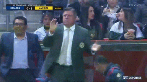 a man in a suit and tie stands in front of a crowd watching a soccer game between rayados and america