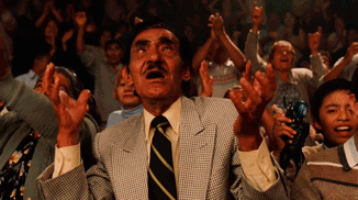 A Man In A Suit And Tie Stands In Front Of A Crowd With His Hands In The Air GIF