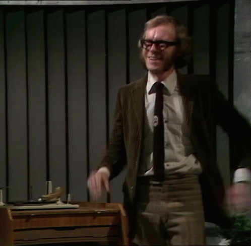 A Man In A Suit And Tie Stands In Front Of A Desk With His Arms Outstretched GIF