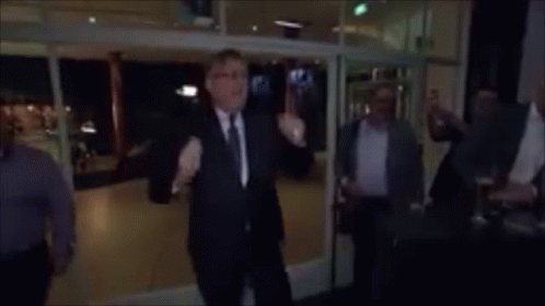A Man In A Suit And Tie Stands In Front Of A Group Of People And Says " Voetjes Van De Vloer " In Yellow Letters GIF