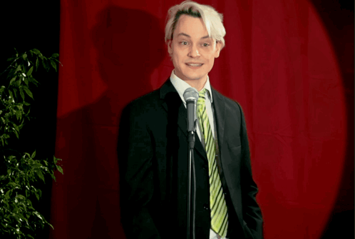 A Man In A Suit And Tie Stands In Front Of A Microphone With The Words " A Bit Desperate " Below Him GIF