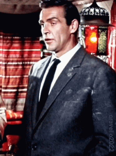 A Man In A Suit And Tie Stands In Front Of A Red Curtain GIF