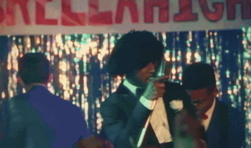 A Man In A Suit And Tie Stands In Front Of A Sign That Says " All-American " GIF